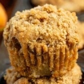 Close-up of an apple muffin topped with golden, crumbly streusel in a dark muffin tin, with another apple muffin and part of an apple blurred in the background.