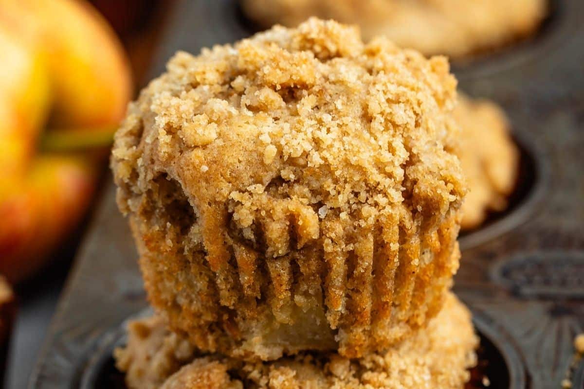 Close-up of an apple muffin topped with golden, crumbly streusel in a dark muffin tin, with another apple muffin and part of an apple blurred in the background.