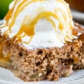 A close-up of a slice of apple pie cake topped with a scoop of vanilla ice cream and drizzled with caramel sauce, served on a white plate.