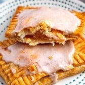Two rectangular apple pop tarts with golden, flaky crusts are stacked on a white plate with blue dots. Topped with white icing, one is broken open to reveal a gooey fruit filling inside.