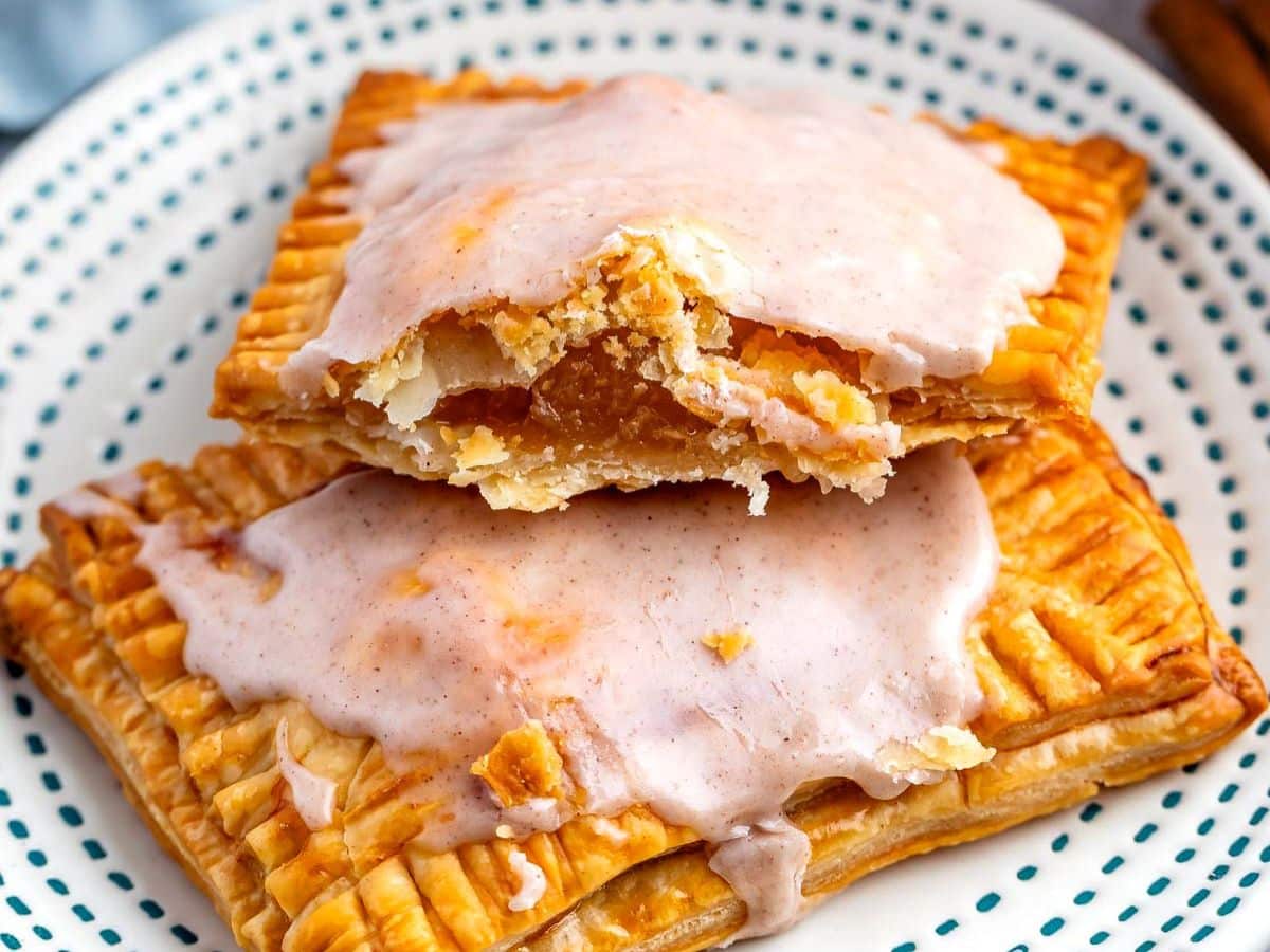 Two rectangular apple pop tarts with golden, flaky crusts are stacked on a white plate with blue dots. Topped with white icing, one is broken open to reveal a gooey fruit filling inside.