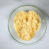 A glass bowl containing a crumbly yellow mixture, likely flour and butter combined to make a crumble topping, sits on a white marble surface.