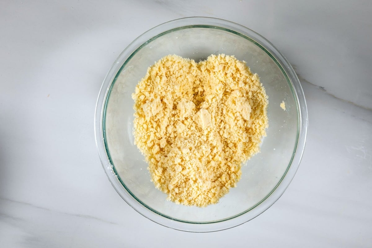 A glass bowl containing a crumbly yellow mixture, likely flour and butter combined to make a crumble topping, sits on a white marble surface.
