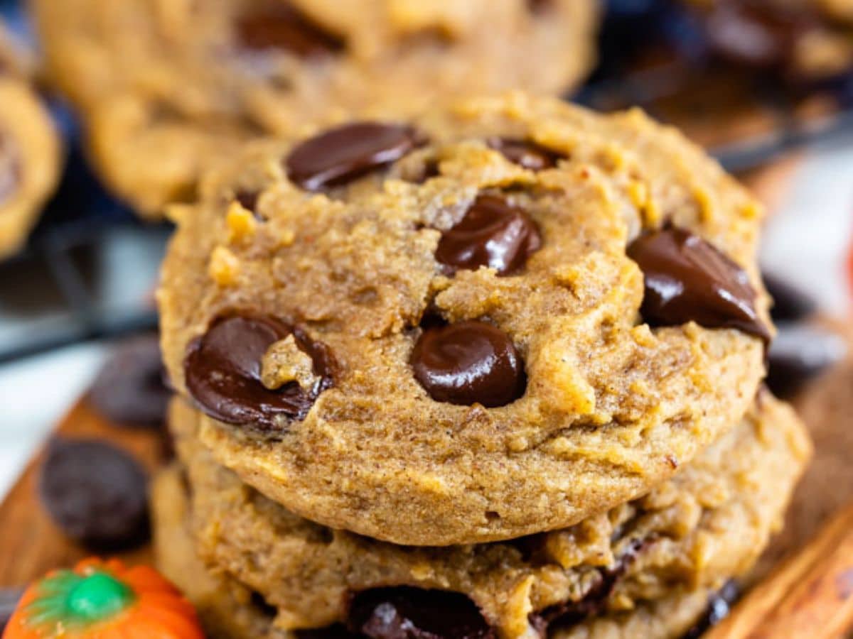 Close-up of two stacked pumpkin chocolate chip cookies on a wooden surface, with melted chocolate chips visible and a small decorative pumpkin nearby.