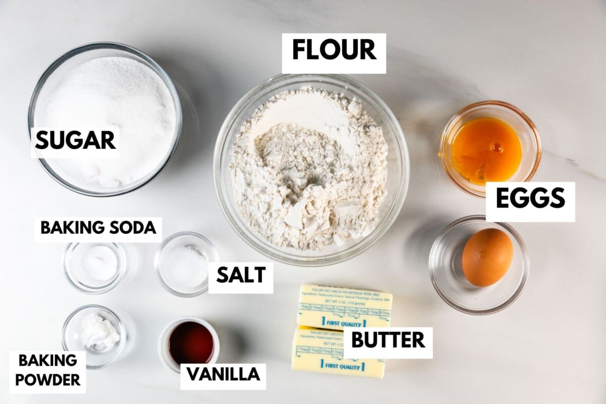 Une vue de haut en bas des ingrédients de cuisson étiquetés pour cette recette facile de gâteau au sucre blanc : deux bols de farine, sucre, œufs, bicarbonate de soude, sel, levure chimique, extrait de vanille et deux bâtonnets de beurre.