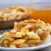 apple pie on a white plate next to a fork and a glass of apple cider.