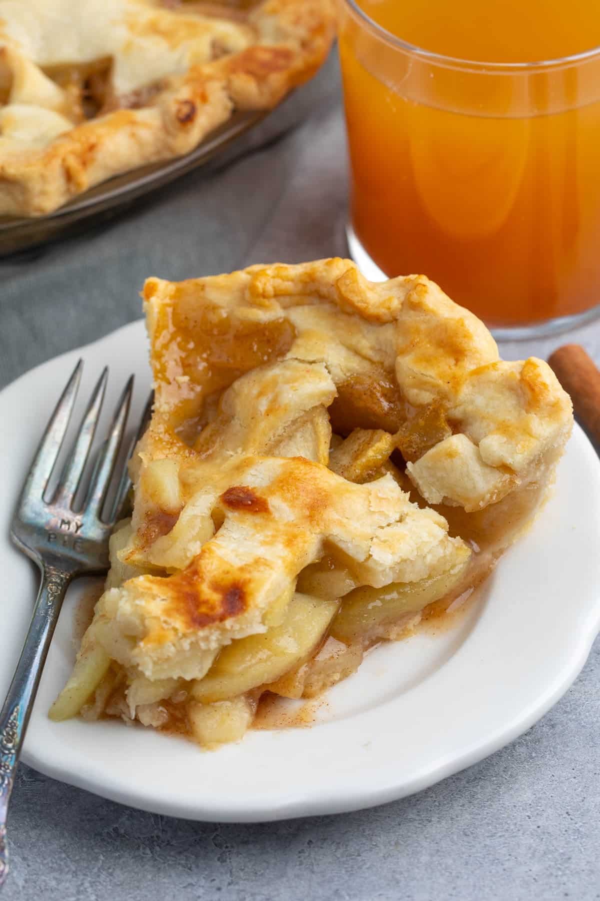 apple pie on a white plate next to a fork and a glass of apple cider.