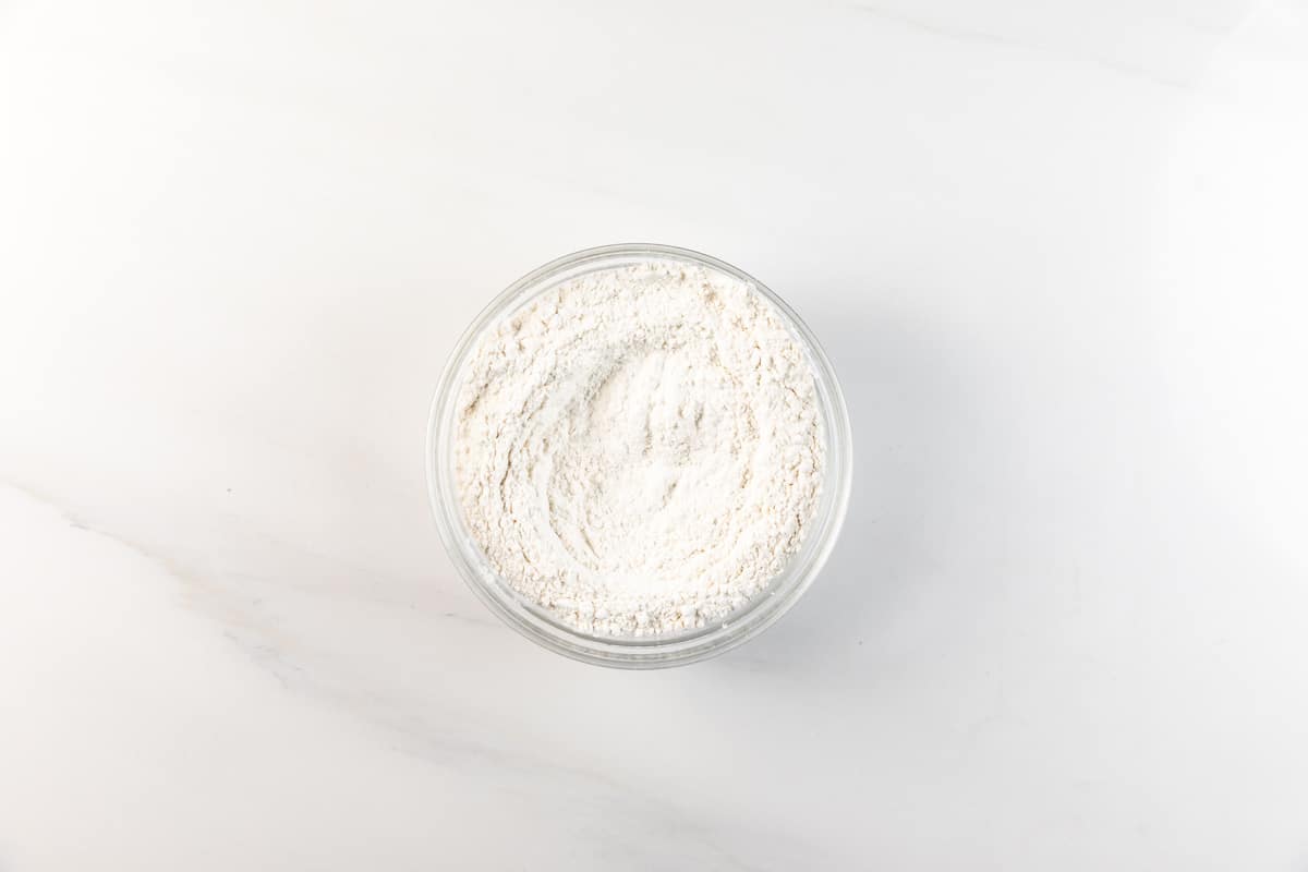 Un bol en verre rempli de farine blanche repose sur une surface en marbre blanc propre – prêt pour votre prochaine recette de gâteau au sucre facile.
