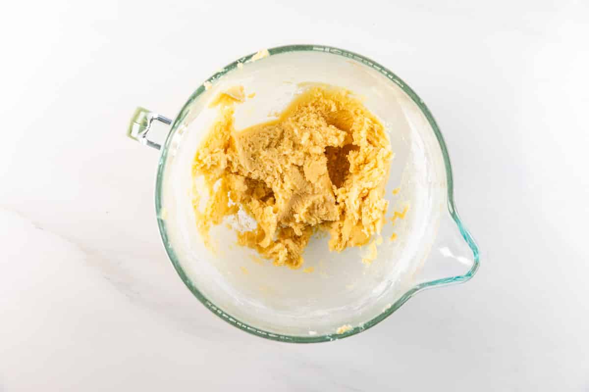 Un bol à mélanger en verre contient une pâte partiellement mélangée pour une recette facile de biscuits au sucre sur une surface blanche. La pâte épaisse et brun clair colle aux parois du bol.
