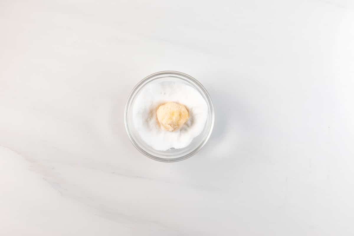 Une petite boule de pâte, prête pour une recette facile de gâteau au sucre, est placée dans un bol en verre transparent rempli de sucre cristallisé sur une surface en marbre blanc.