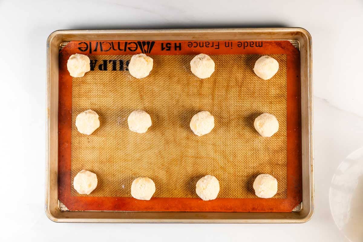 Une plaque à pâtisserie recouverte d'un tapis en silicone contient douze boules de pâte uniformément espacées provenant d'une recette facile de pain au sucre, prêtes à cuire.