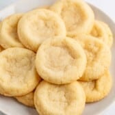 A plate of round, golden sugar cookies made from an easy sugar cookie recipe, with slightly cracked surfaces and soft centers, arranged in a pile on a white plate.