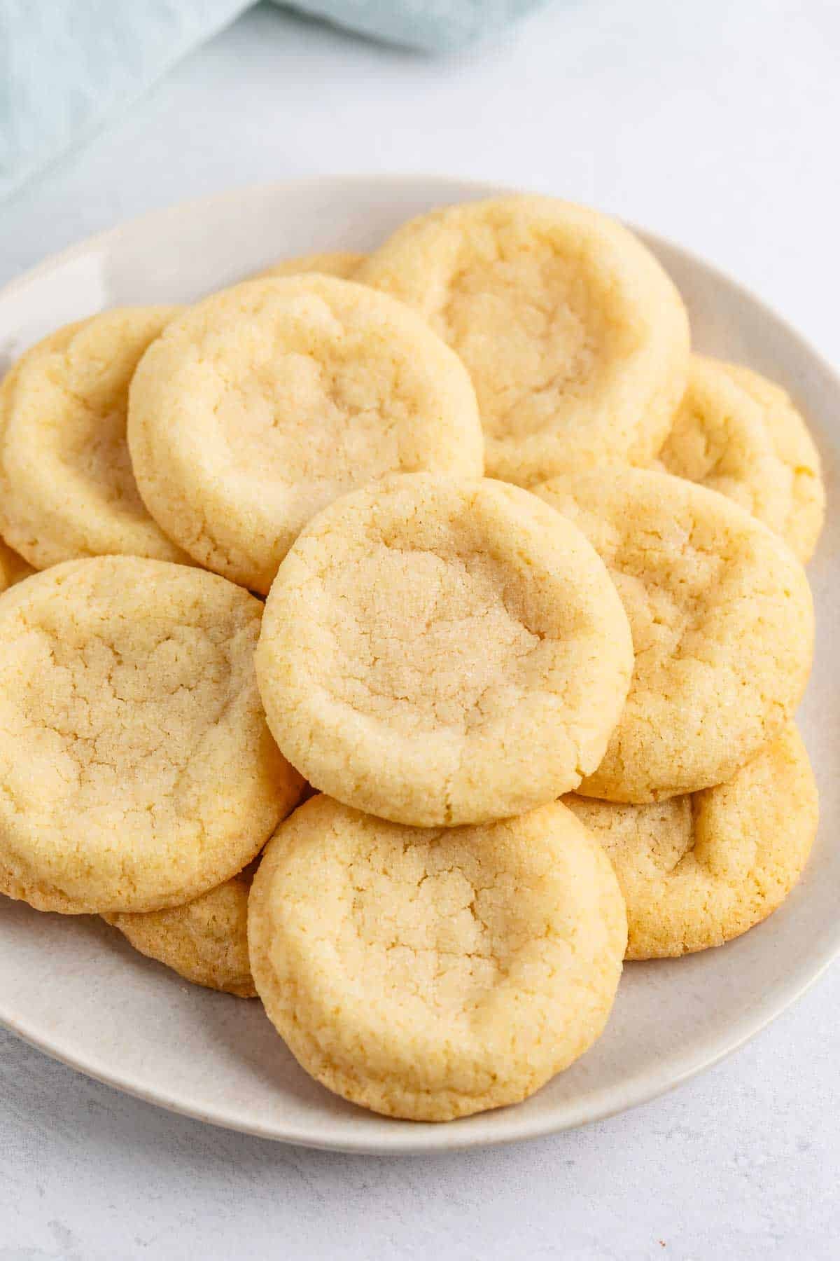 A plate of round, golden sugar cookies made from an easy sugar cookie recipe, with slightly cracked surfaces and soft centers, arranged in a pile on a white plate.