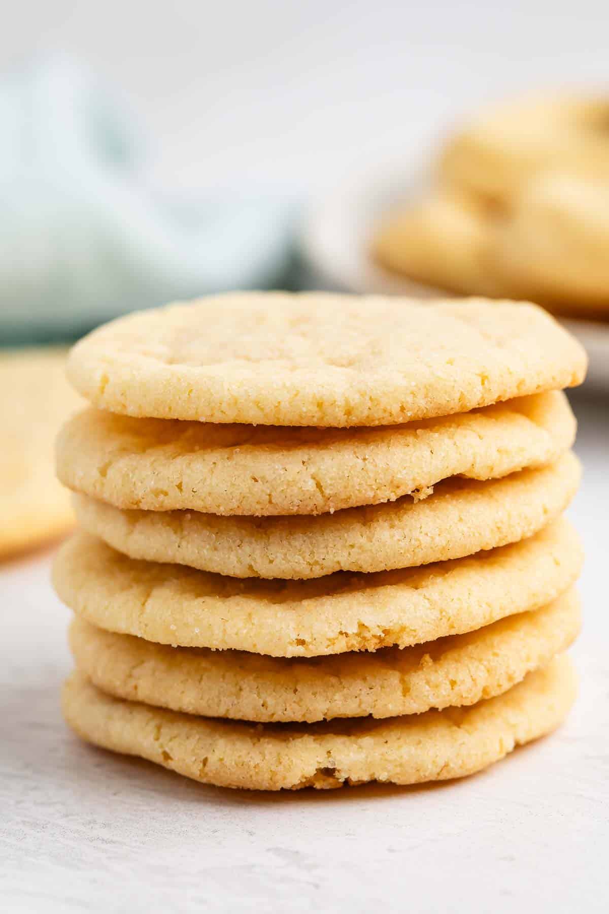 Une pile de six biscuits ronds et dorés, fabriqués à partir d'une recette de biscuits facile, disposés sur une surface claire, avec d'autres biscuits flous en arrière-plan. Les biscuits semblent lisses avec des bords légèrement prononcés.
