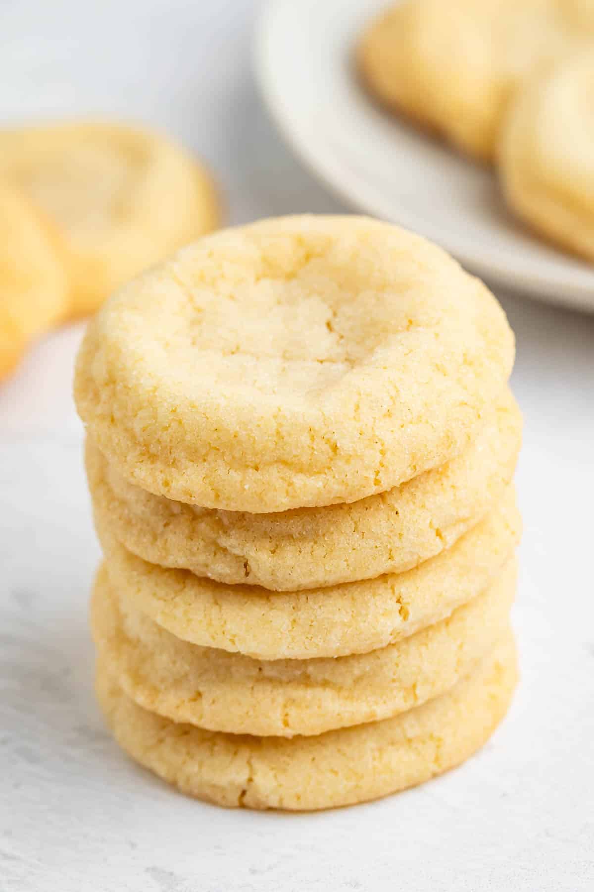 Une pile de cinq biscuits au sucre doré repose sur une surface claire, cuite à la perfection selon une recette facile de biscuits au sucre, avec d'autres biscuits flous en arrière-plan sur une assiette blanche.
