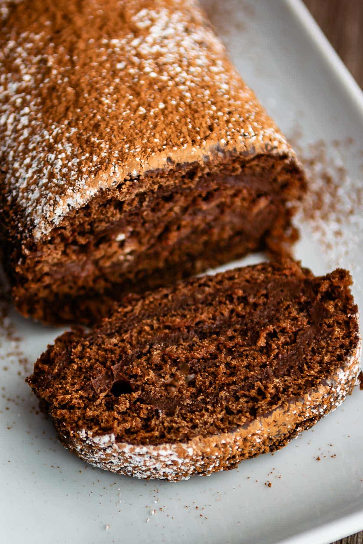 brown cake roll on a white plate with brown and white sugar sprinkled over the top of the roll.