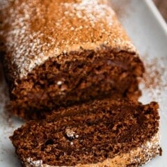 brown cake roll on a white plate with brown and white sugar sprinkled over the top of the roll.