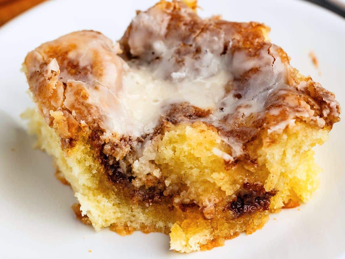 A close-up of a Cinnamon Roll Cake slice with swirls of cinnamon sugar, topped with creamy white glaze, served on a white plate.