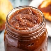 A close-up of a glass jar filled with thick, brown crock pot apple butter. In the background, there is a whole apple and a slice of bread spread with the rich apple butter on a white plate.