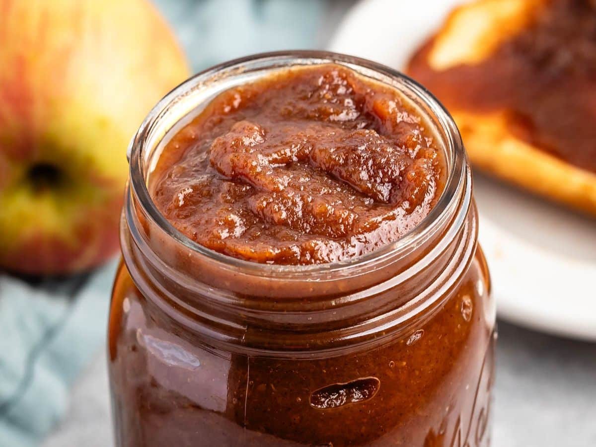 A close-up of a glass jar filled with thick, brown crock pot apple butter. In the background, there is a whole apple and a slice of bread spread with the rich apple butter on a white plate.