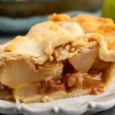 A close-up of a slice of pear pie with a golden, flaky crust and cinnamon-spiced pear filling on a white plate.