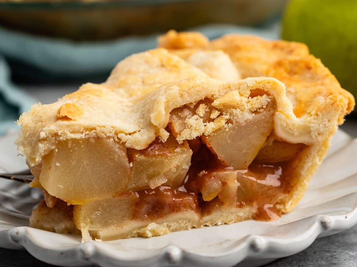 A close-up of a slice of pear pie with a golden, flaky crust and cinnamon-spiced pear filling on a white plate.
