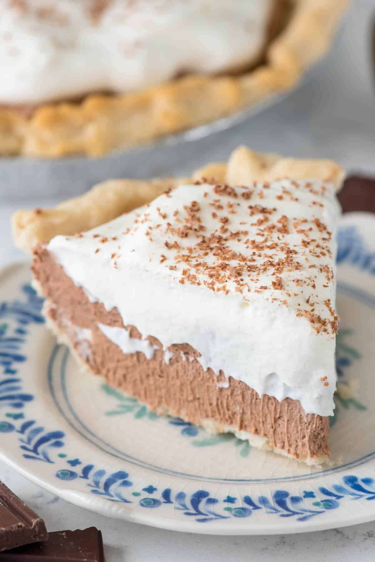 A slice of rich chocolate cream pie topped with whipped cream and chocolate shavings sits on a decorative plate, while the rest of the chocolate cream pie is visible in the background.