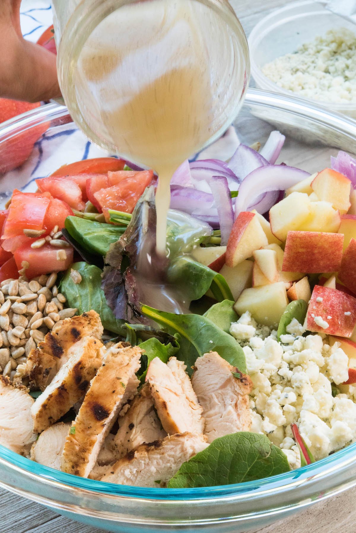 salad mixed with apples and onions and chicken and more in a clear bowl with a dressing being poured in.