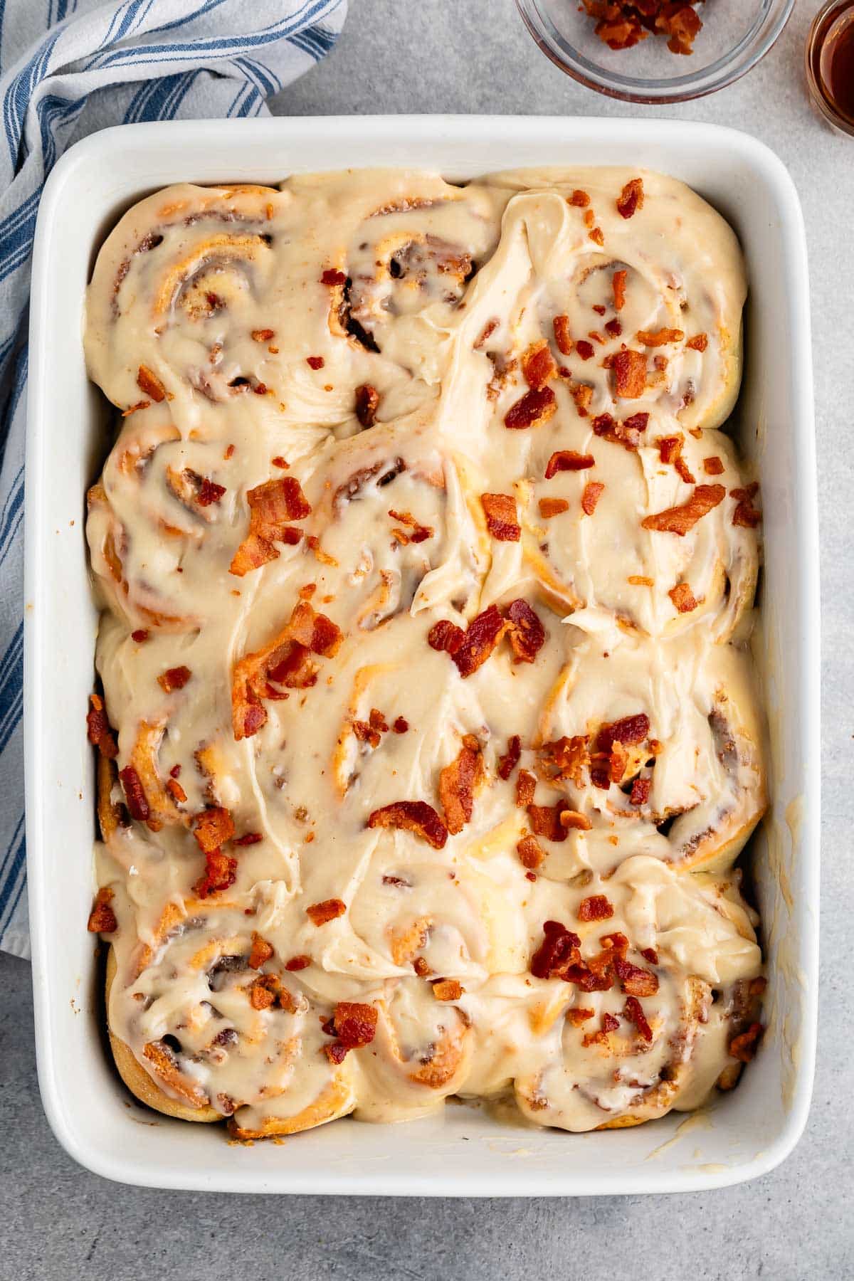 maple bacon cinnamon roll iced with white icing and sprinkled with bacon in a white dish.