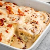 maple bacon cinnamon roll iced with white icing and sprinkled with bacon in a white dish.