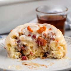 maple bacon cinnamon roll iced with white icing and sprinkled with bacon in a white dish.