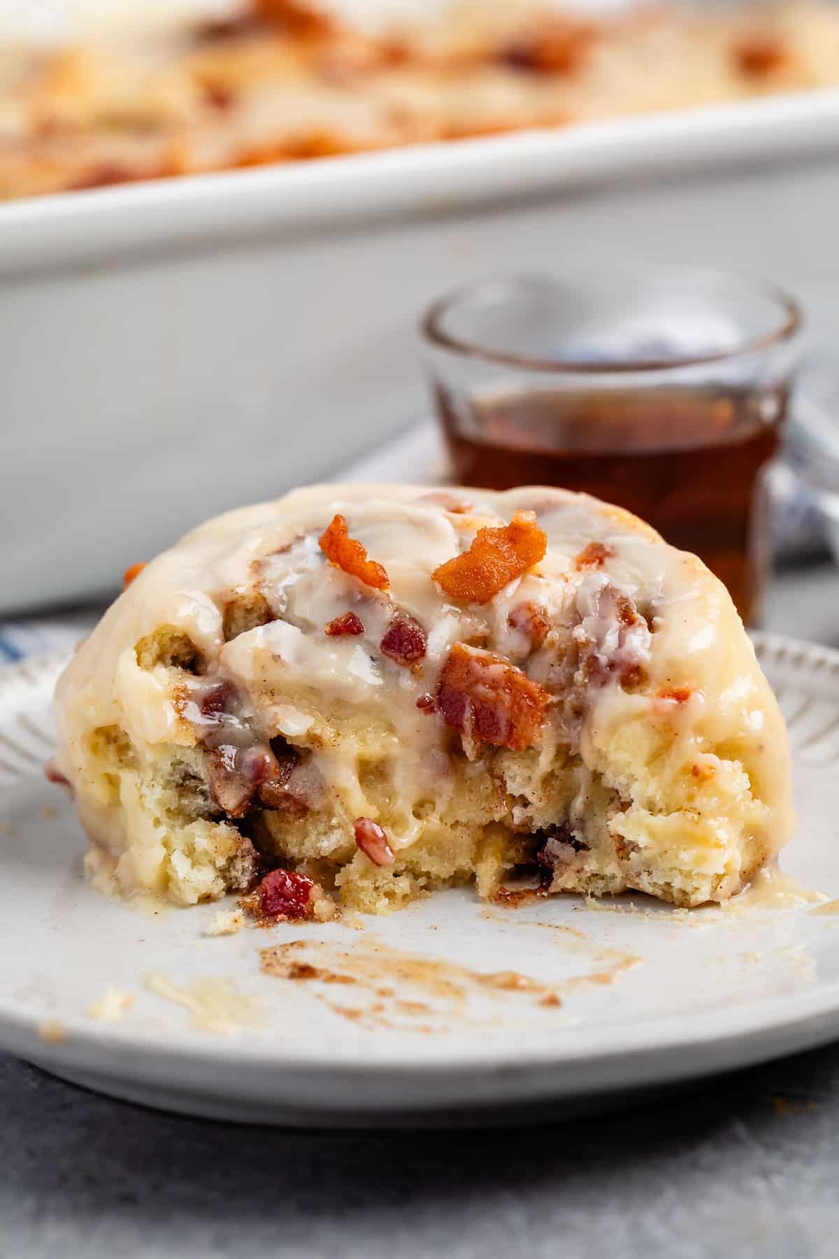 maple bacon cinnamon roll iced with white icing and sprinkled with bacon in a white dish.