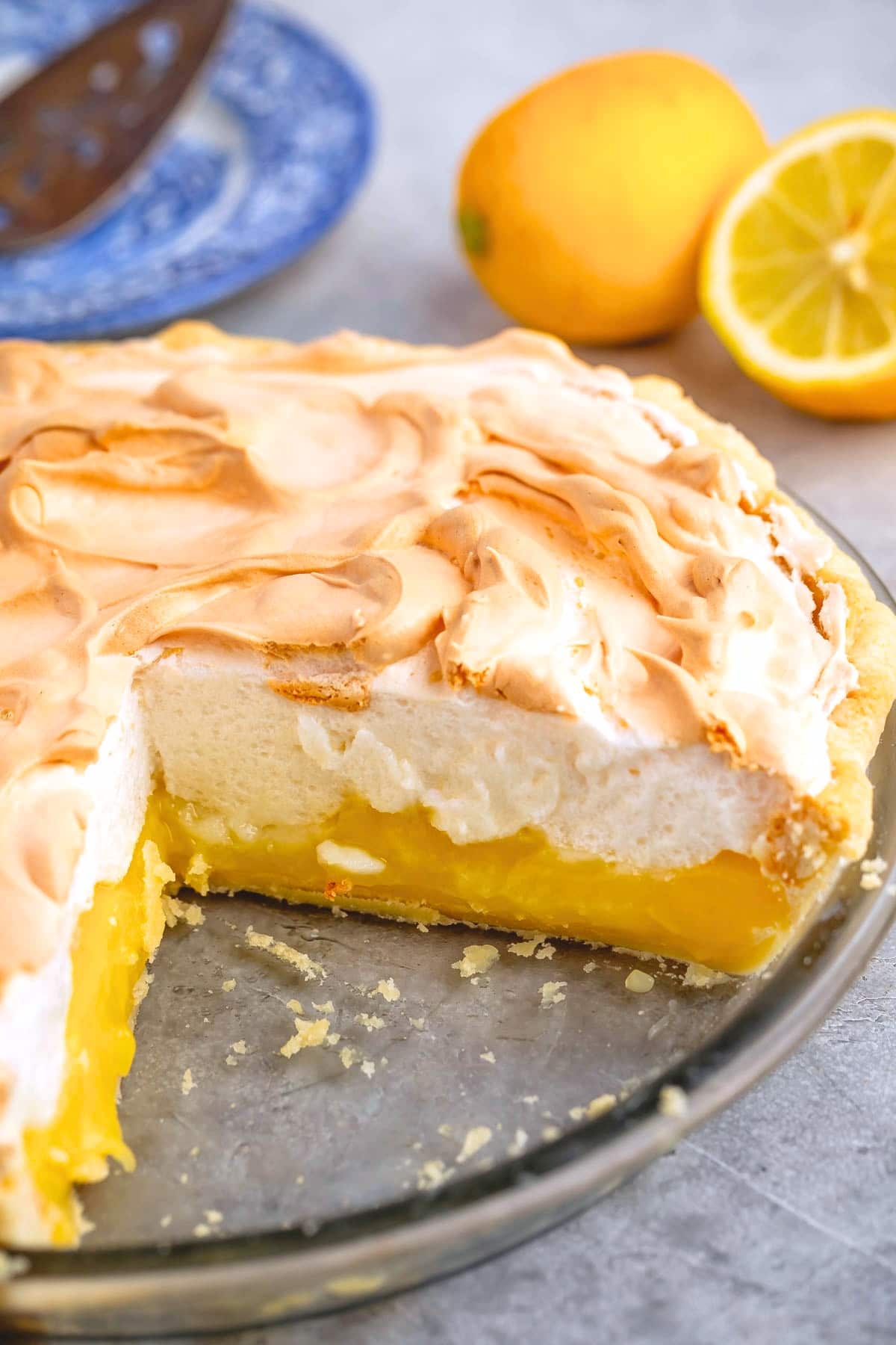 A lemon meringue pie with a slice missing, showing layers of vibrant yellow lemon filling and fluffy meringue topping. A halved lemon and a whole lemon sit in the background on a gray surface, completing this classic lemon meringue pie scene.