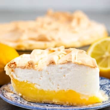 A slice of lemon meringue pie sits on a blue-patterned plate, with a whole lemon, a lemon half, and the rest of the delicious lemon meringue pie in the background. The pie features a vibrant yellow lemon layer and a fluffy meringue topping.
