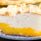A slice of lemon meringue pie sits on a blue patterned plate, showing off its vibrant yellow lemon filling topped with fluffy, lightly browned meringue. In the background, a cut lemon hints at the zesty flavors in this classic dessert.