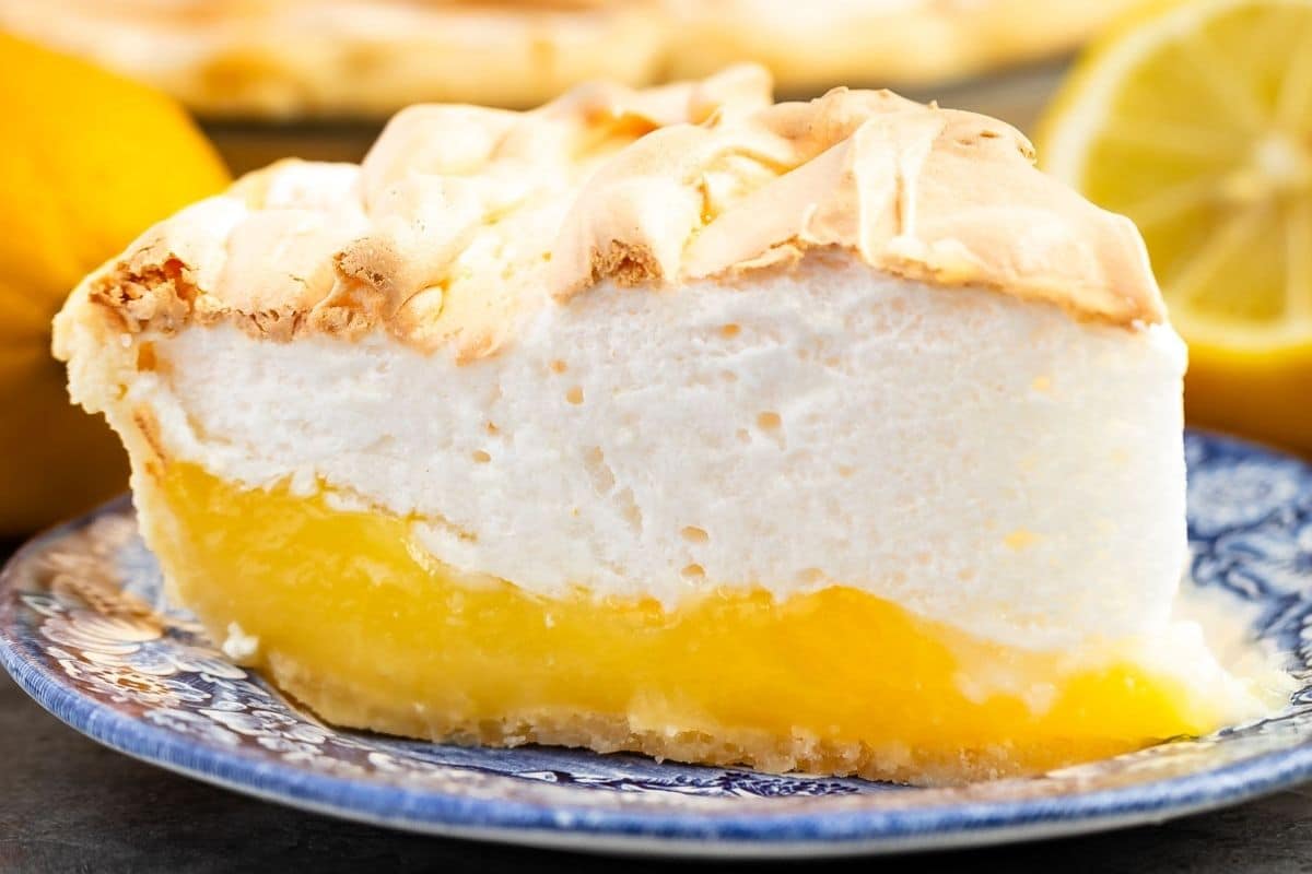 A slice of lemon meringue pie sits on a blue patterned plate, showing off its vibrant yellow lemon filling topped with fluffy, lightly browned meringue. In the background, a cut lemon hints at the zesty flavors in this classic dessert.