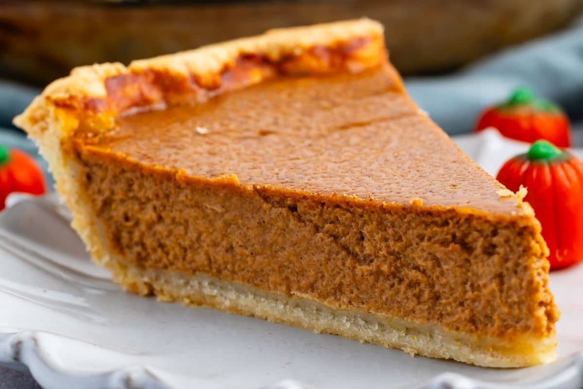 A close-up of a slice of pumpkin pie on a white plate, with a thick, creamy orange filling and a golden crust—just like the classic results you’d get from your favorite pumpkin pie recipe. Small decorative pumpkin-shaped candies are in the background.