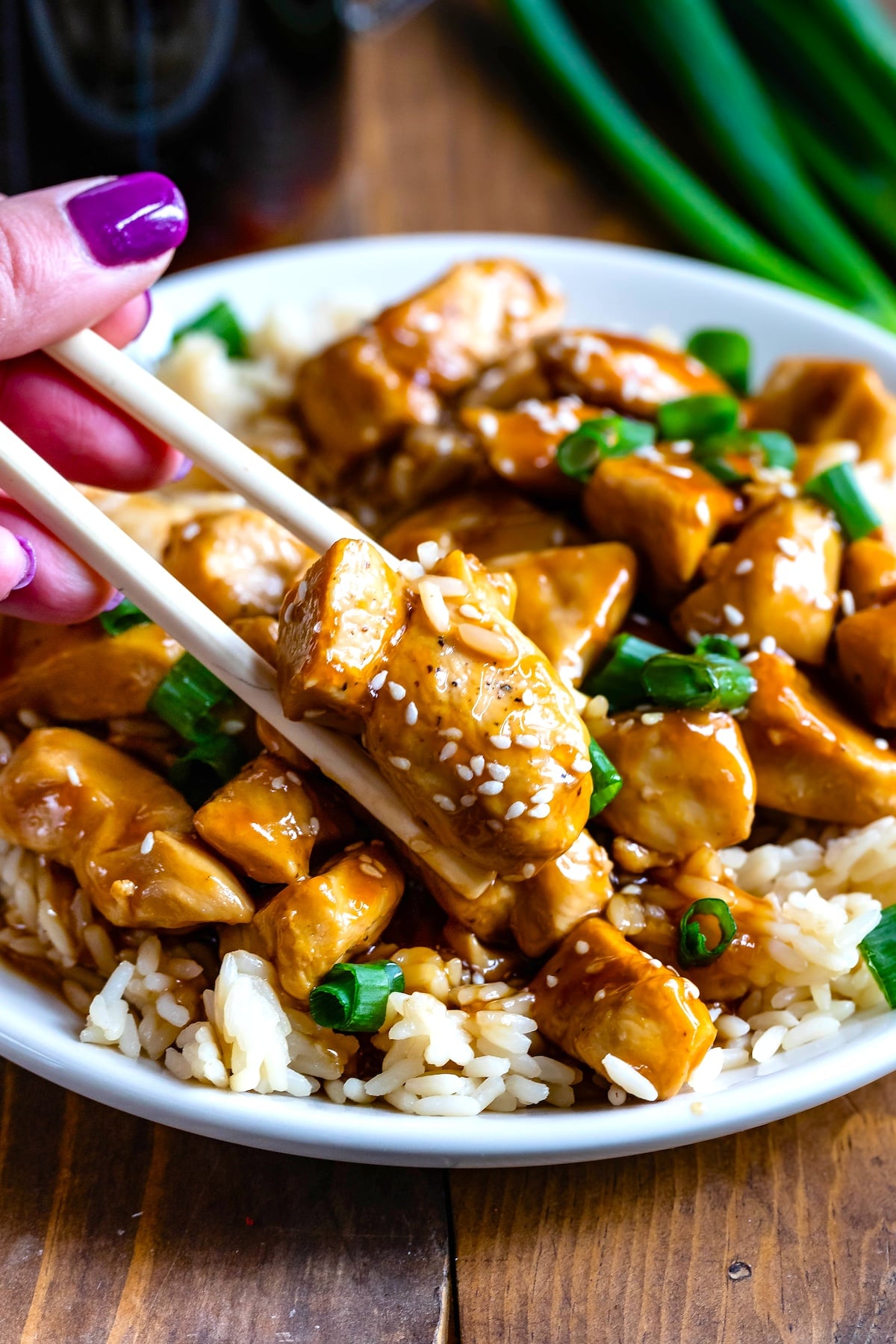 hand picking up teriyaki chicken with chopsticks on top of rice on a white plate with teriyaki sauce in the background.