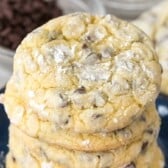 A stack of freshly baked Cake Mix Crinkle Cookies dusted with powdered sugar sits on a blue plate, with a bowl of chocolate chips visible in the background.