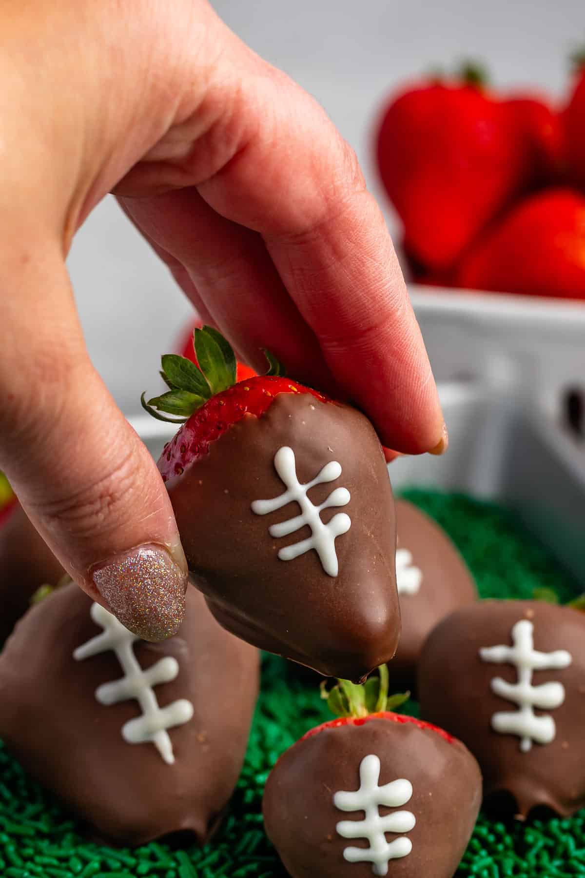 hand holding chocolate covered strawberries with white laces drawn on top with green sprinkles underneath.