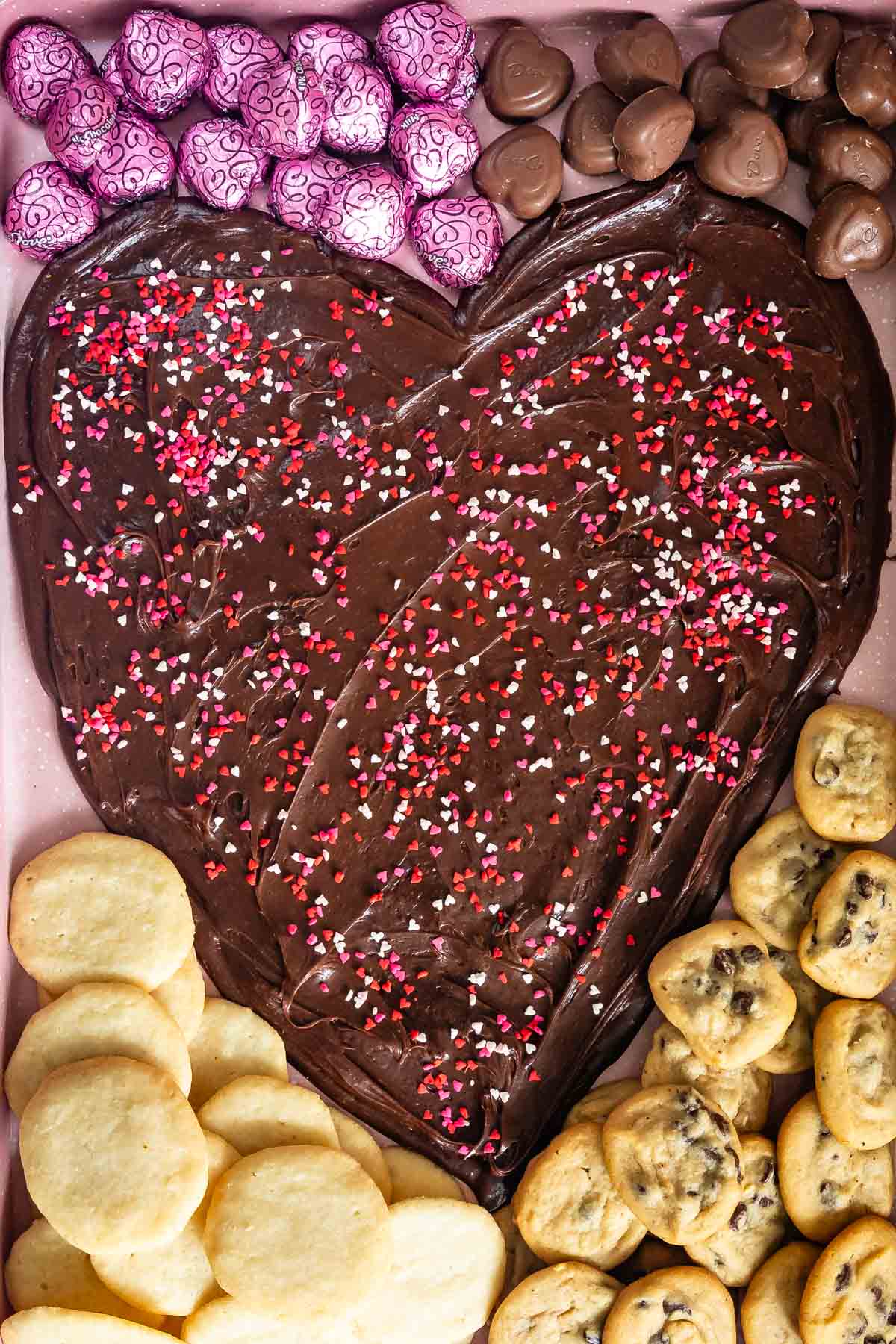 nutella spread on a pink board in the shape of a heart with candy and cookies around the edge.