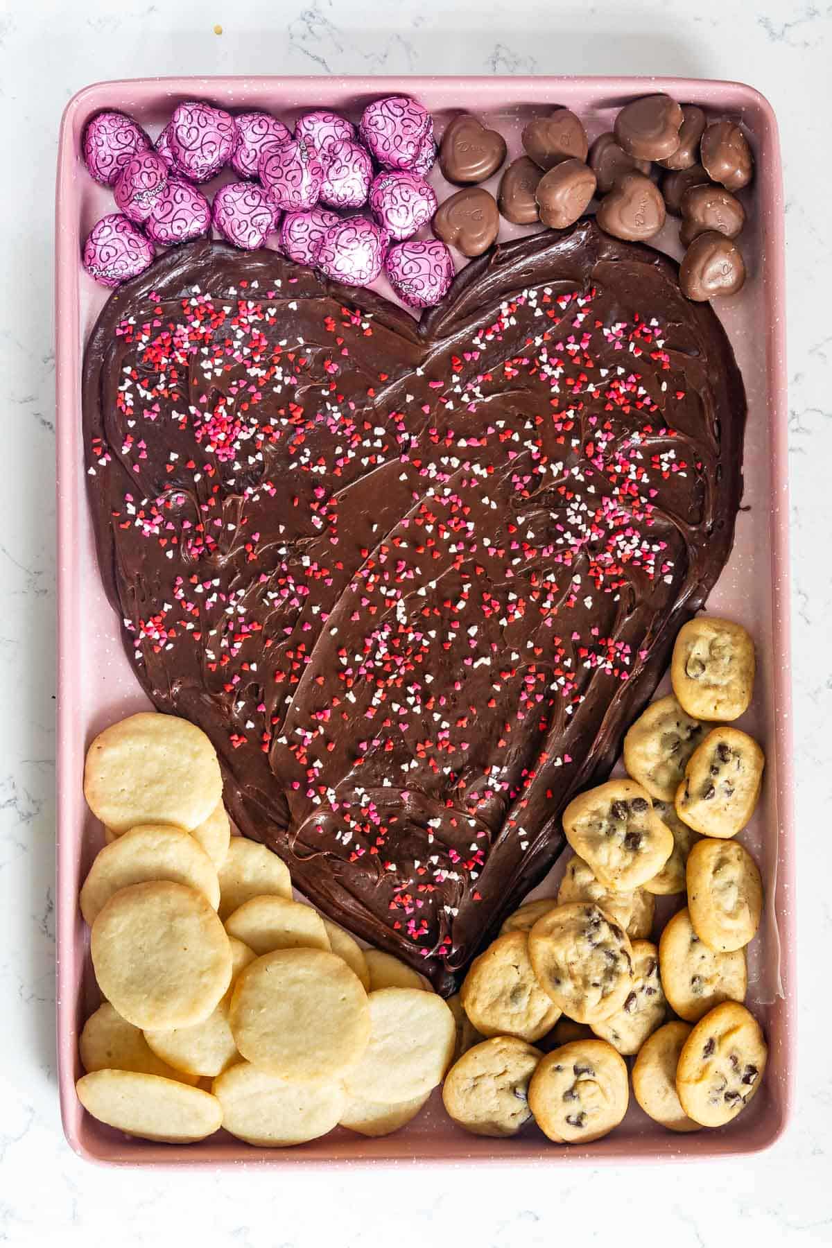 nutella spread on a pink board in the shape of a heart with candy and cookies around the edge.