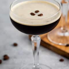 espresso martini in a tall clear glass with coffee beans all around the glass.