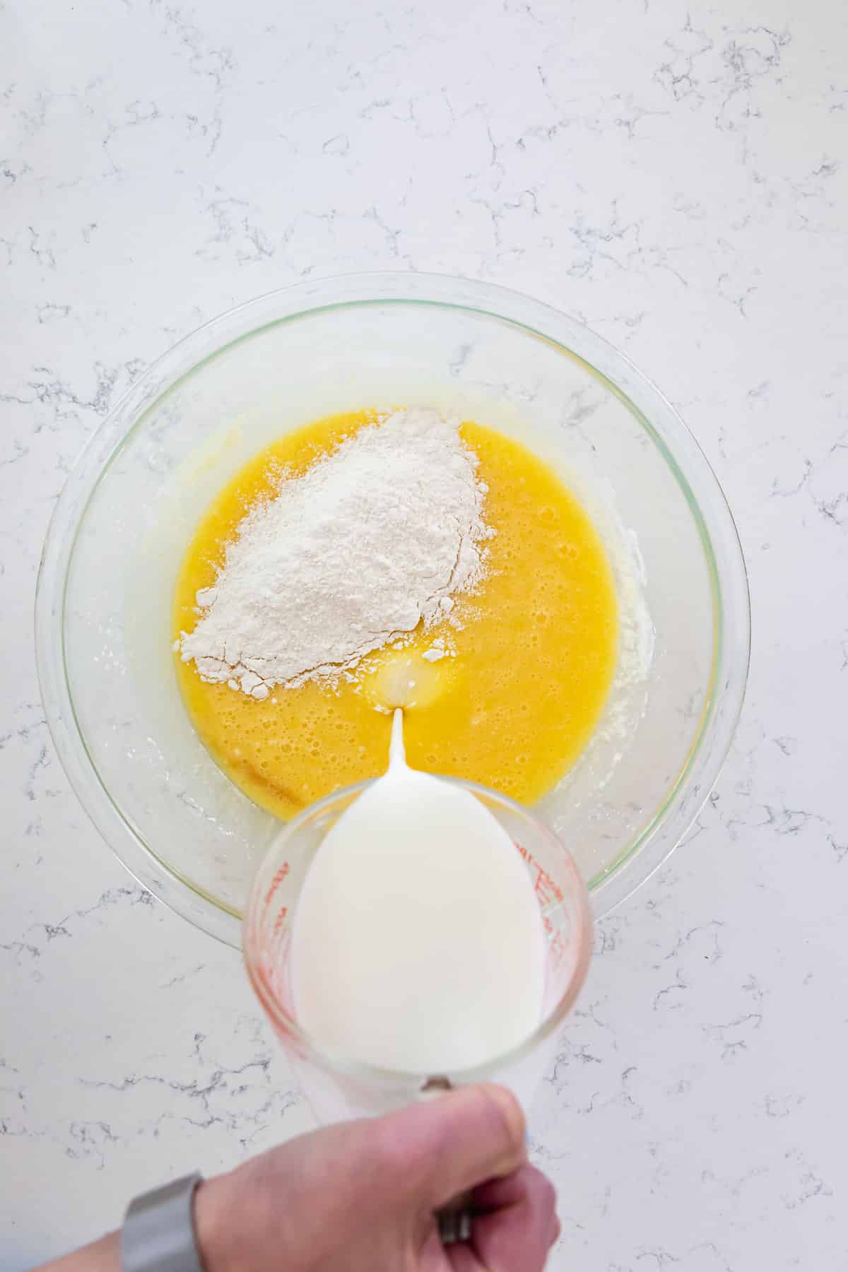cake batter in glass bowl with flour and hand pouring milk into batter on marble background.