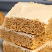 Three decadent Peanut Butter Bars stacked on a white plate, showcasing their thick, golden-brown texture and creamy frosting on top. The background is softly blurred with a hint of peanut butter visible, highlighting the irresistible appeal of these classic bars.