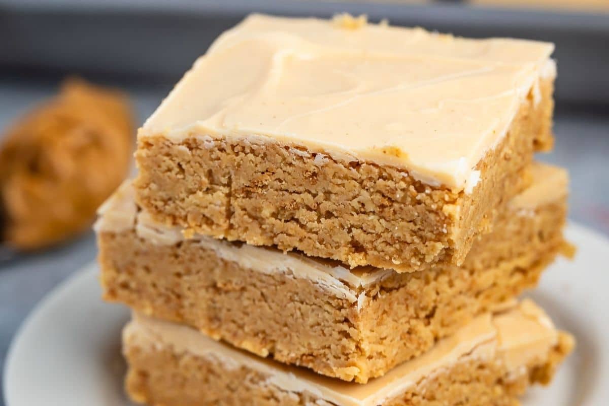 Three decadent Peanut Butter Bars stacked on a white plate, showcasing their thick, golden-brown texture and creamy frosting on top. The background is softly blurred with a hint of peanut butter visible, highlighting the irresistible appeal of these classic bars.