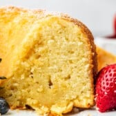 A close-up of a slice of moist, golden bundt cake made from an easy pound cake recipe, lightly dusted with powdered sugar and surrounded by fresh strawberries and blueberries on a white plate.