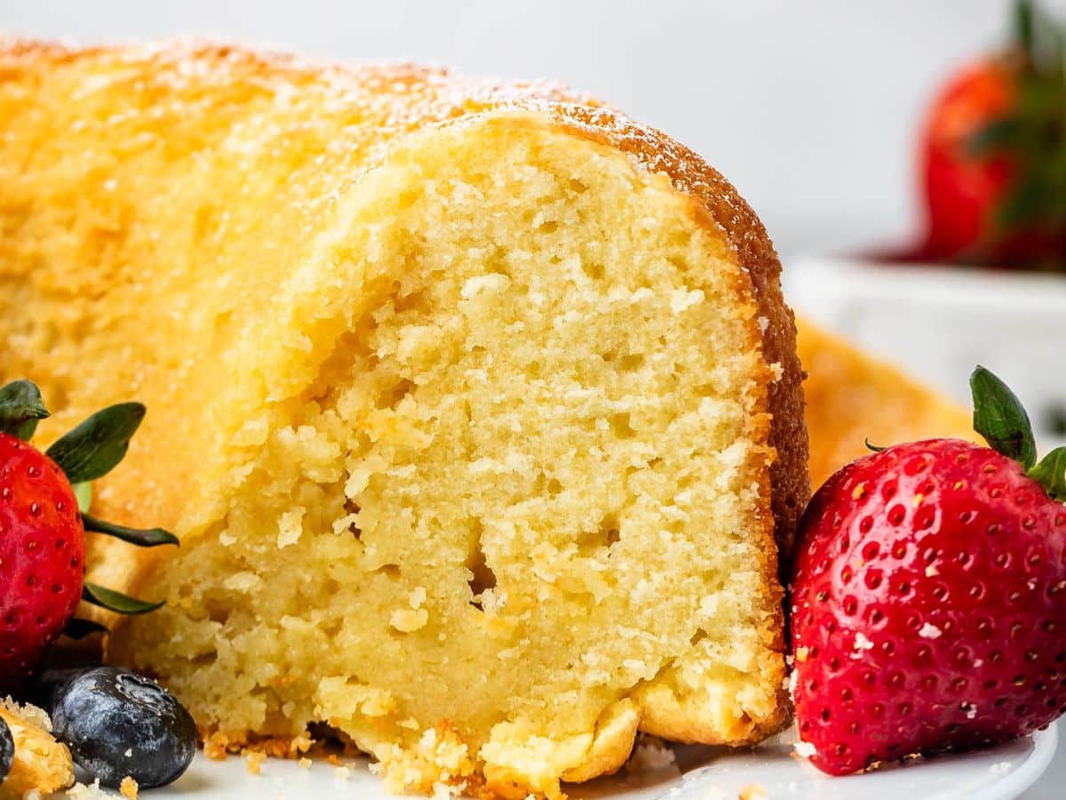 A close-up of a slice of moist, golden bundt cake made from an easy pound cake recipe, lightly dusted with powdered sugar and surrounded by fresh strawberries and blueberries on a white plate.