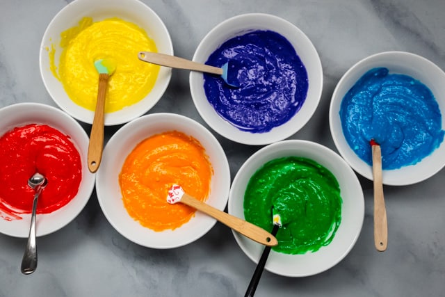 rainbow colors of cake batter in white bowls with spatulas.