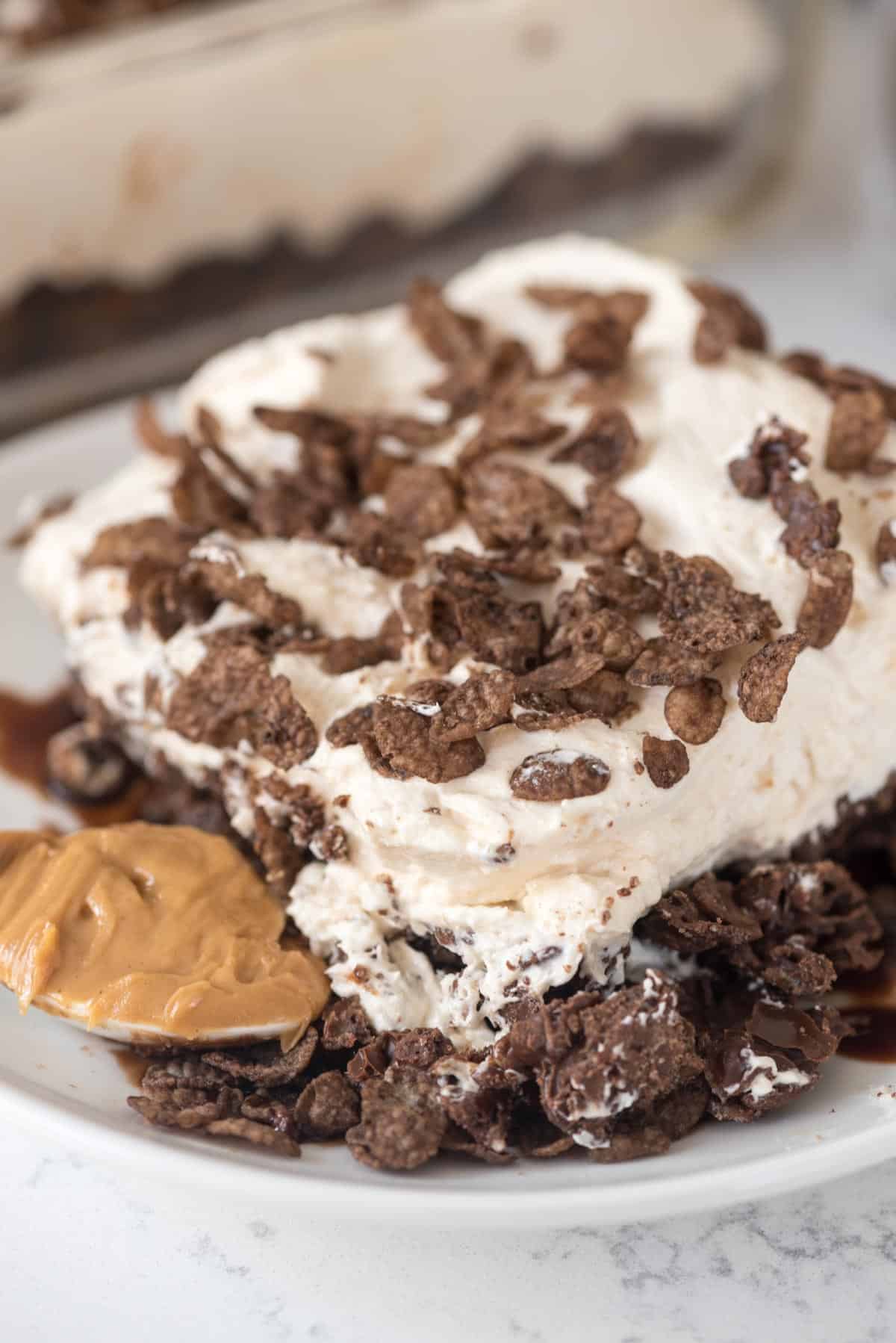 cocoa pebble dessert layered with chocolate and cream on a white plate next to a spoonful of peanut butter.